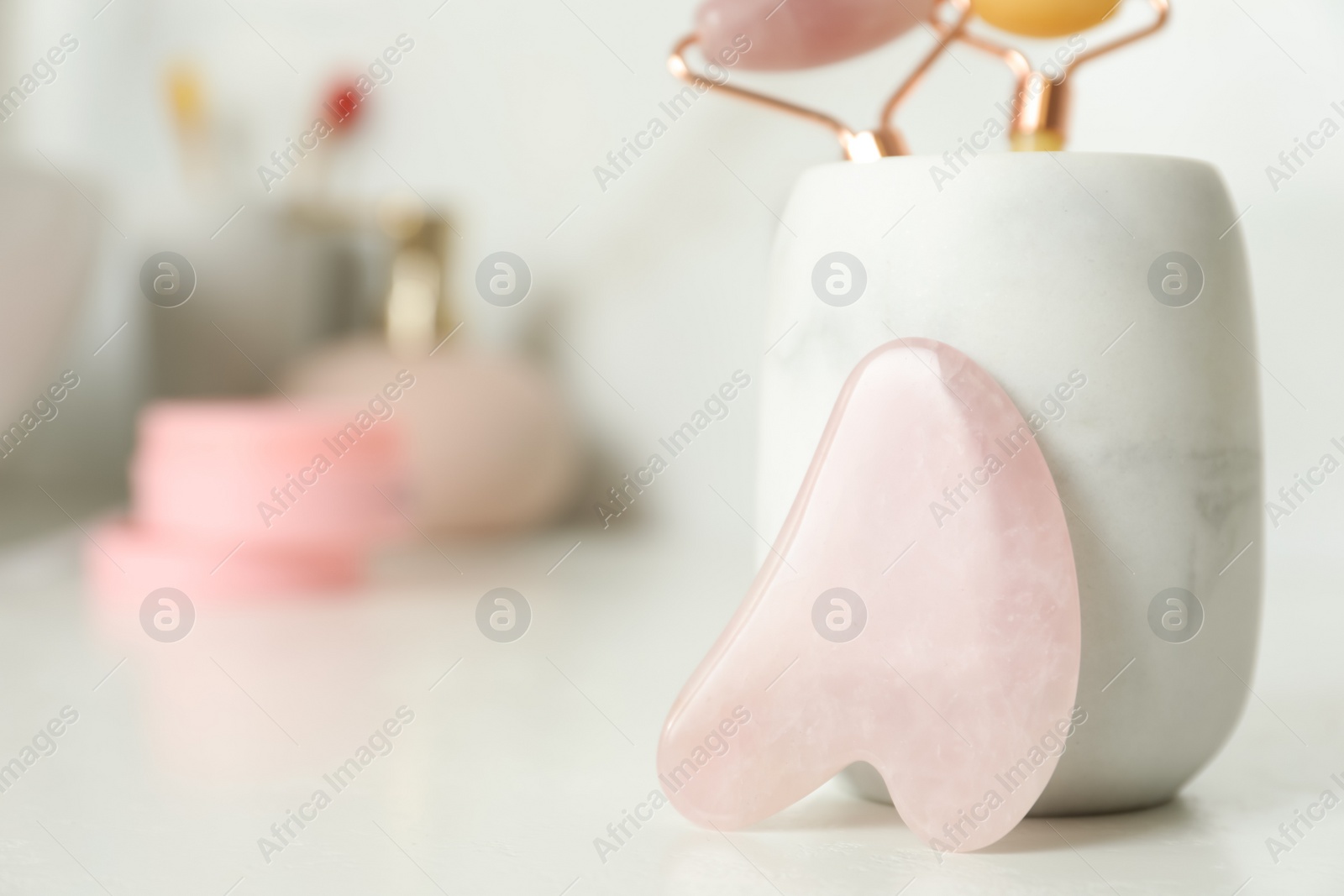 Photo of Rose quartz gua sha tool near holder with natural face rollers on white countertop in bathroom, closeup. Space for text