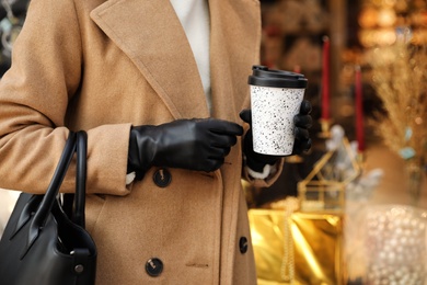 Woman with stylish leather gloves and cup of coffee near store decorated for Christmas, closeup