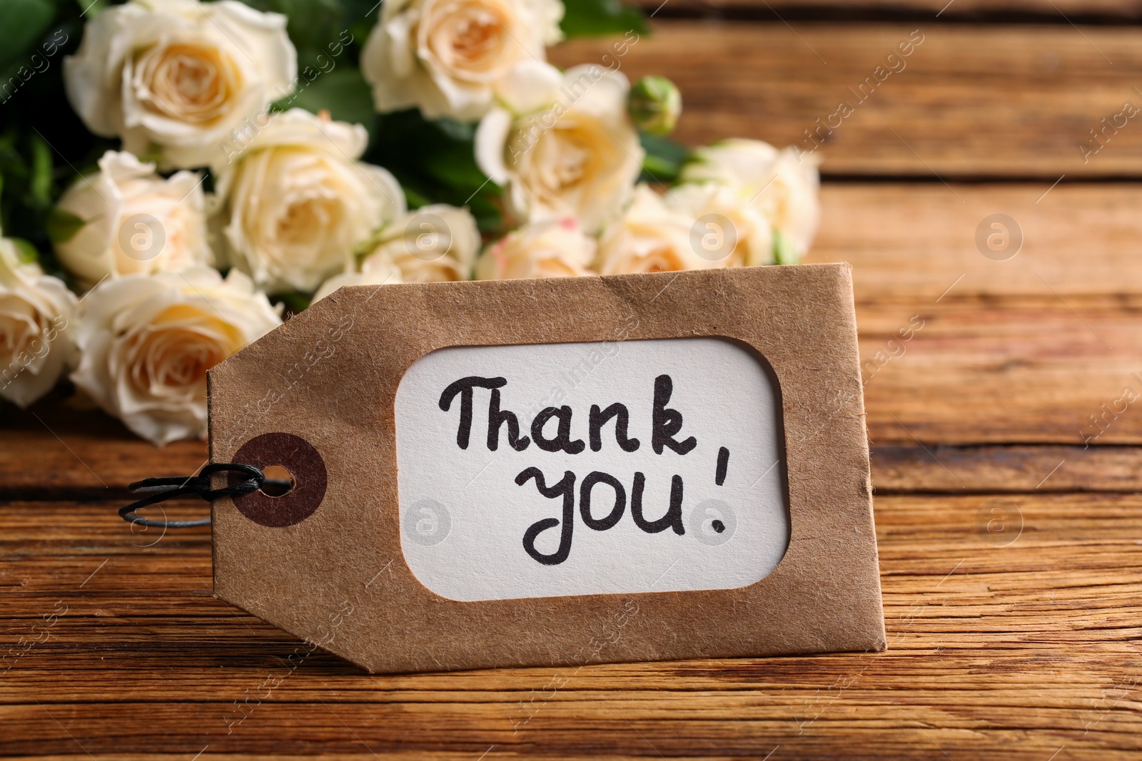 Photo of Tag with phrase Thank you and roses on wooden table, closeup