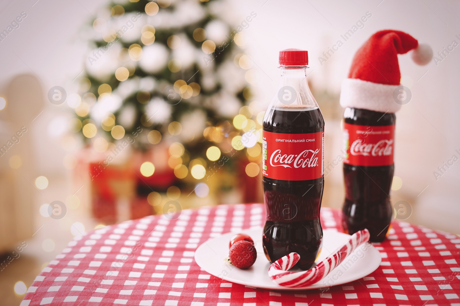 Photo of MYKOLAIV, UKRAINE - January 01, 2021: Bottles of Coca-Cola and candy cane on table against blurred Christmas lights