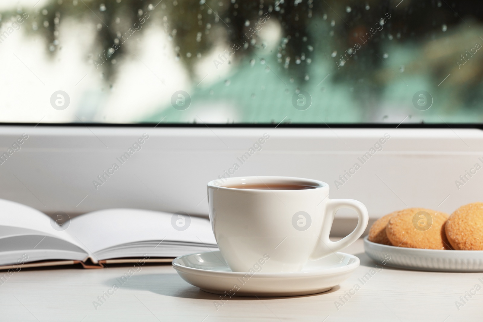 Photo of Cup of tea, saucer with cookies and open book on wooden window sill