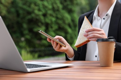 Businesswoman with sandwich using smartphone while having lunch at wooden table outdoors, closeup