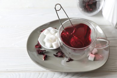 Delicious hibiscus tea in cup, sugar cubes and dry roselle petals on white wooden table, above view. Space for text