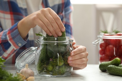 Woman putting cucumber into glass jar at white marble kitchen table, closeup. Pickling vegetables