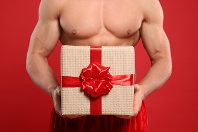 Young man with muscular body holding Christmas gift box on red background, closeup