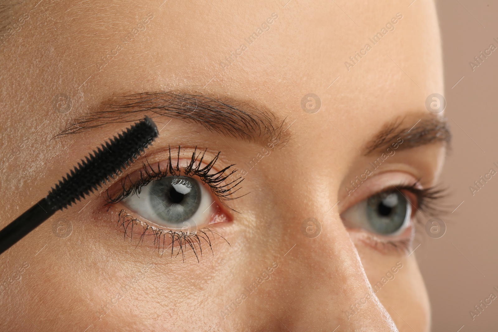 Photo of Woman applying mascara onto eyelashes against light brown background, closeup