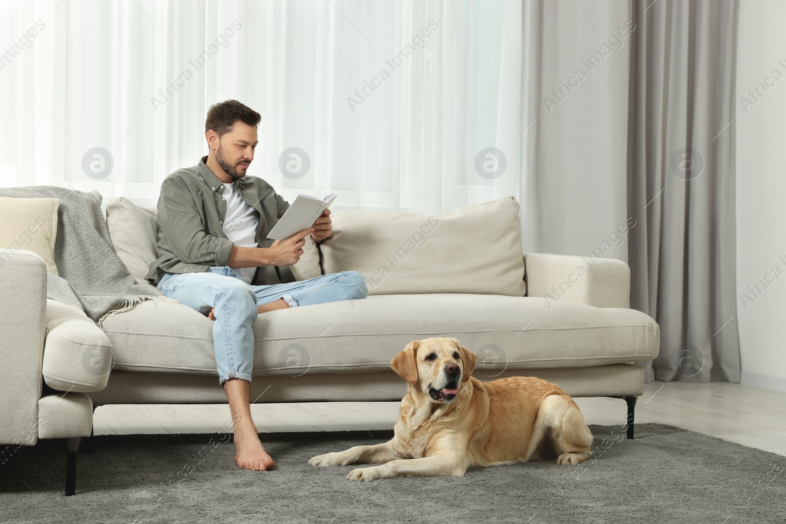 Photo of Man reading book on sofa near his cute Labrador Retriever at home