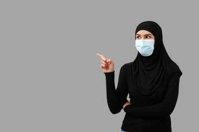Photo of Muslim woman in hijab and medical mask pointing at something on light gray background, space for text