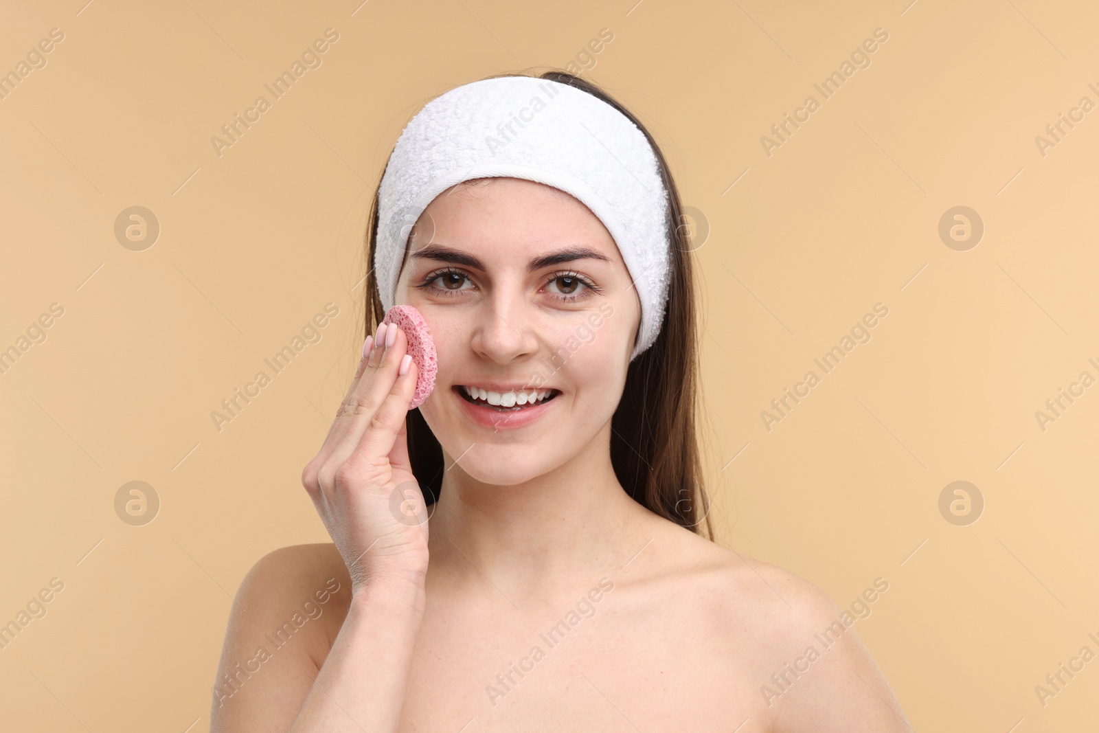 Photo of Young woman with headband washing her face using sponge on beige background