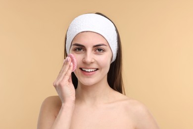 Photo of Young woman with headband washing her face using sponge on beige background