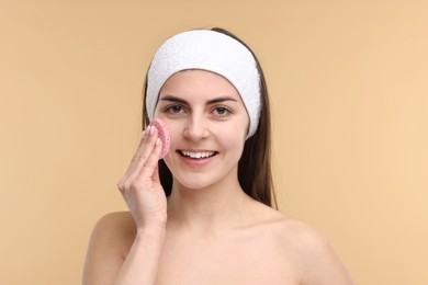 Young woman with headband washing her face using sponge on beige background