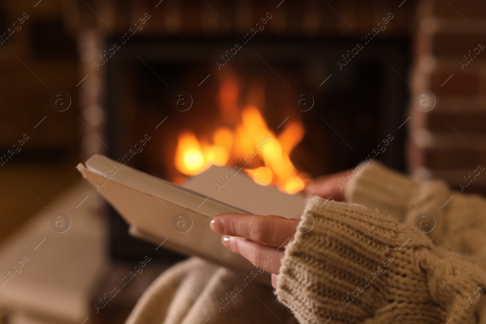 Photo of Woman reading book near burning fireplace at home, closeup