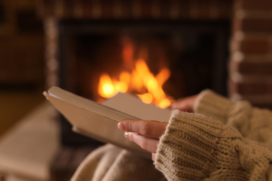 Photo of Woman reading book near burning fireplace at home, closeup