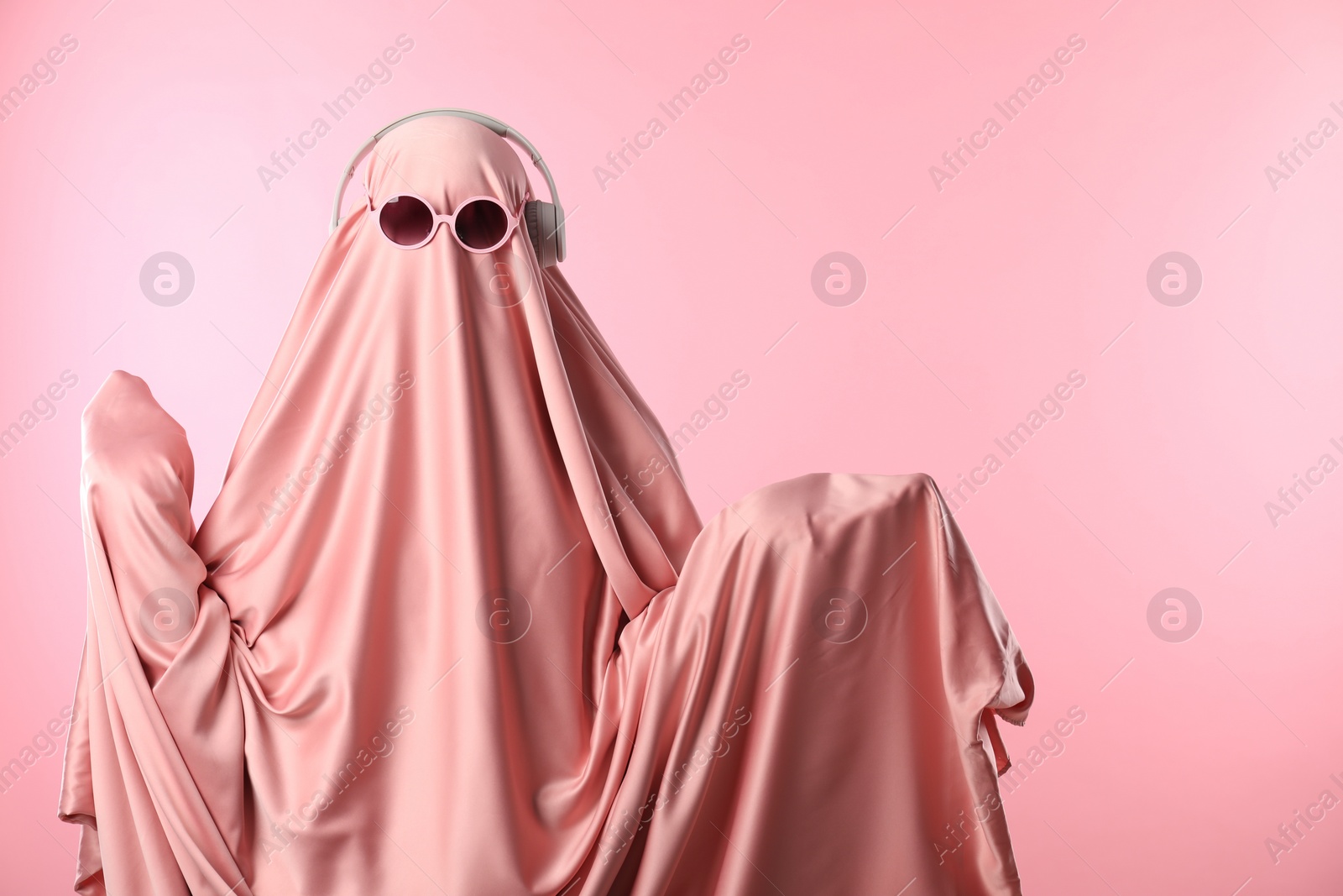 Photo of Glamorous ghost. Woman in sheet with sunglasses and headphones on pink background, space for text