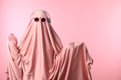 Glamorous ghost. Woman in sheet with sunglasses and headphones on pink background, space for text