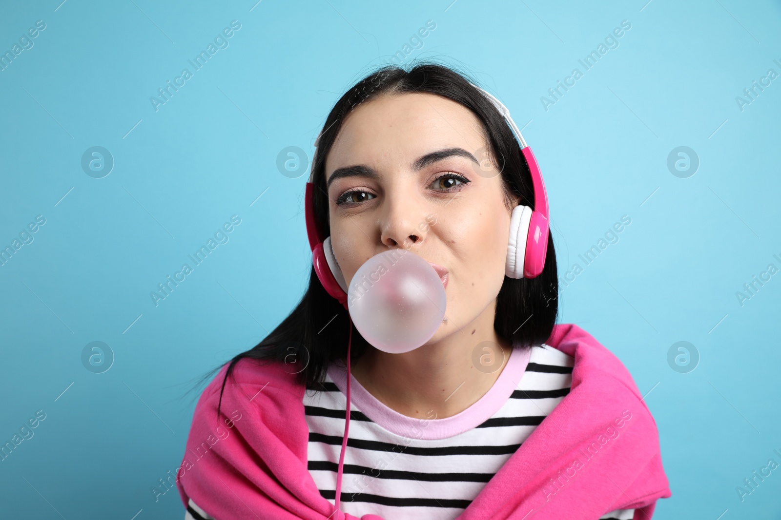 Photo of Fashionable young woman with headphones blowing bubblegum on light blue background