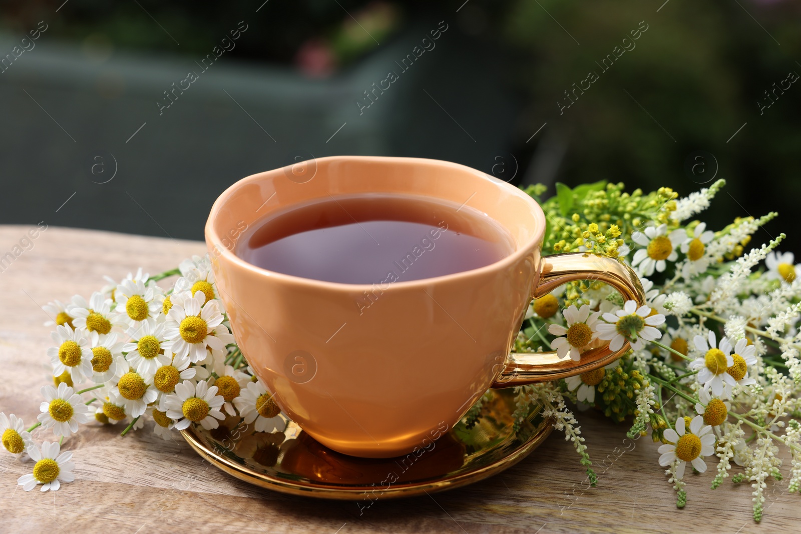 Photo of Cup of delicious chamomile tea and fresh flowers outdoors, closeup