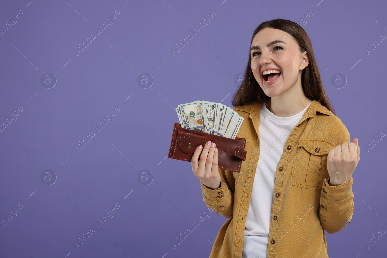 Photo of Excited woman holding wallet with dollar banknotes on purple background, space for text