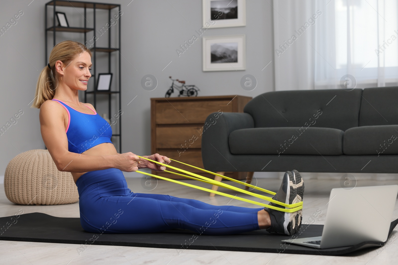 Photo of Fit woman doing exercise with fitness elastic band near laptop on mat at home