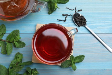 Photo of Cup of aromatic black tea with mint and dry leaves on light blue wooden table, flat lay