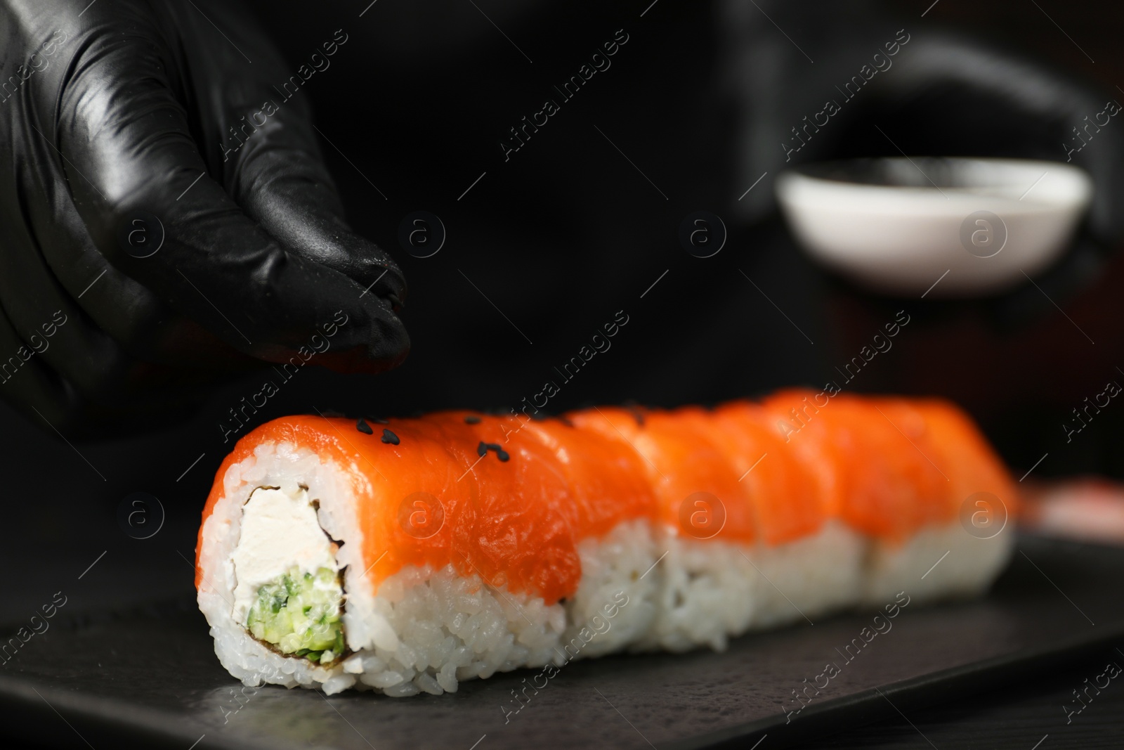 Photo of Chef in gloves adding sesame seeds onto tasty sushi rolls at table, closeup