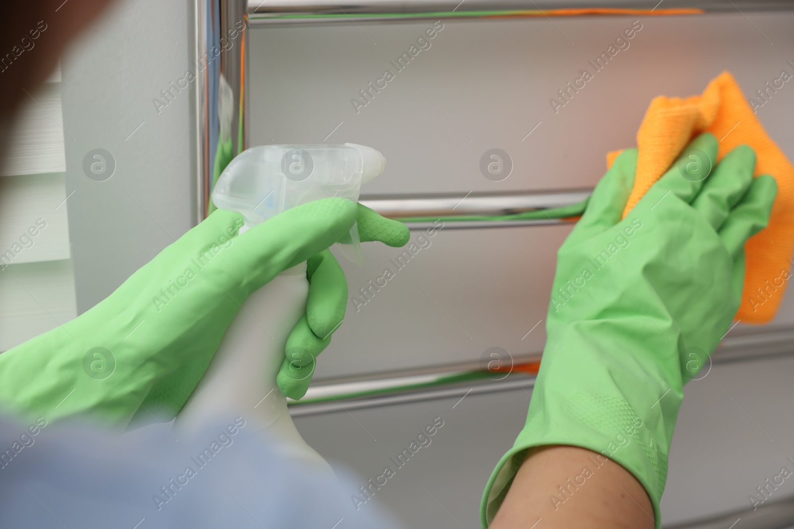 Photo of Woman cleaning heated towel rail with sprayer and rag, closeup