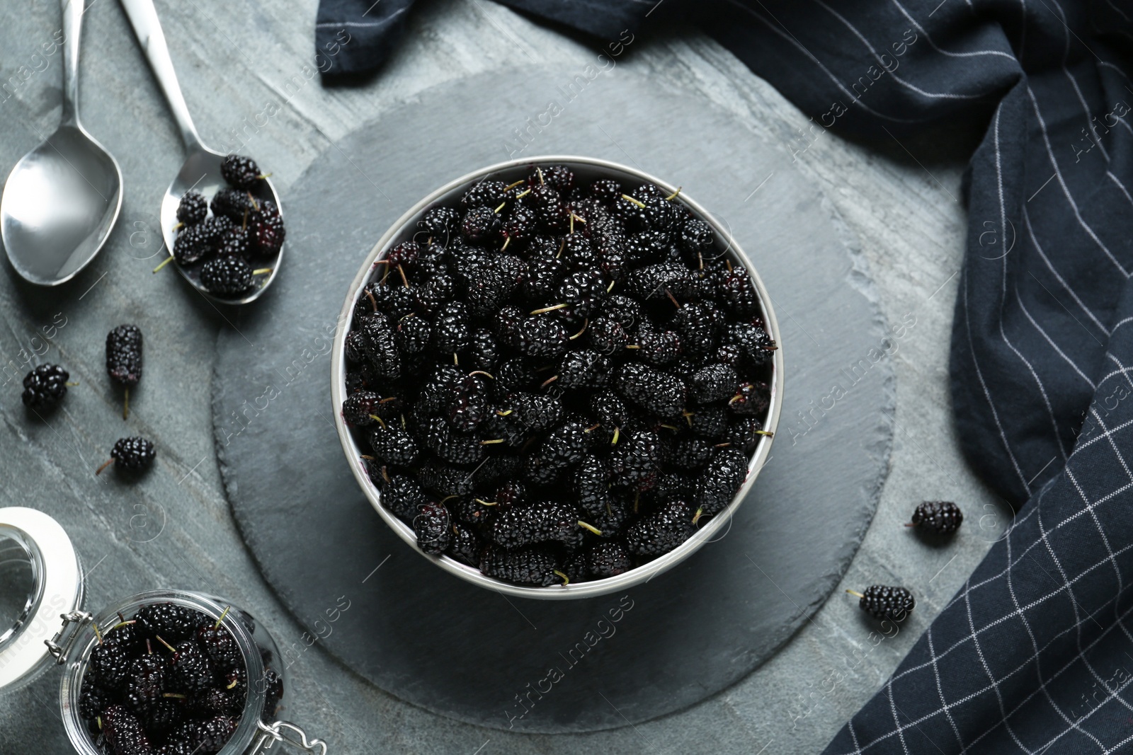 Photo of Bowl, spoon and jar of delicious ripe black mulberries on grey table, flat lay