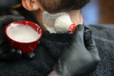 Professional hairdresser applying shaving foam onto client's beard in barbershop, closeup