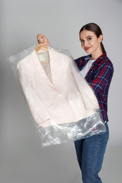 Young woman holding hanger with jacket on light grey background. Dry-cleaning service