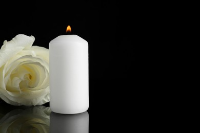Photo of White rose and burning candle on black mirror surface in darkness, closeup with space for text. Funeral symbols