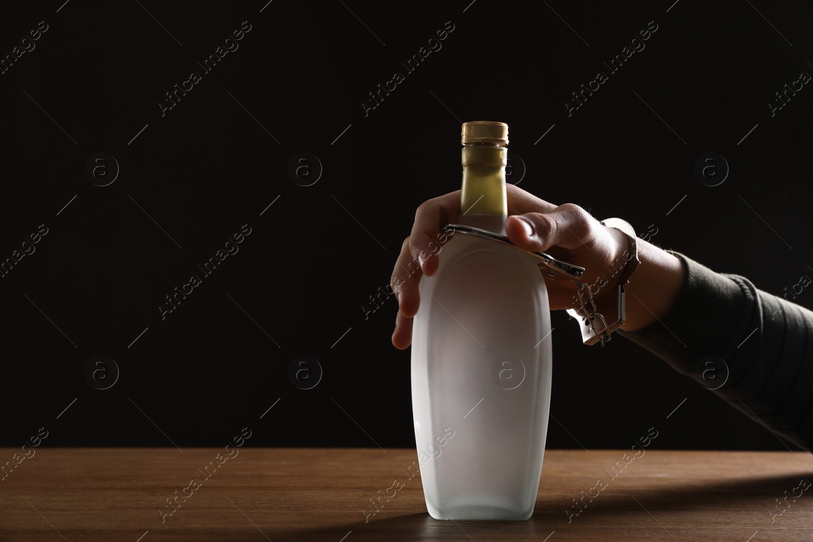 Photo of Alcohol addiction. Man handcuffed to bottle of vodka at wooden table, closeup. Space for text