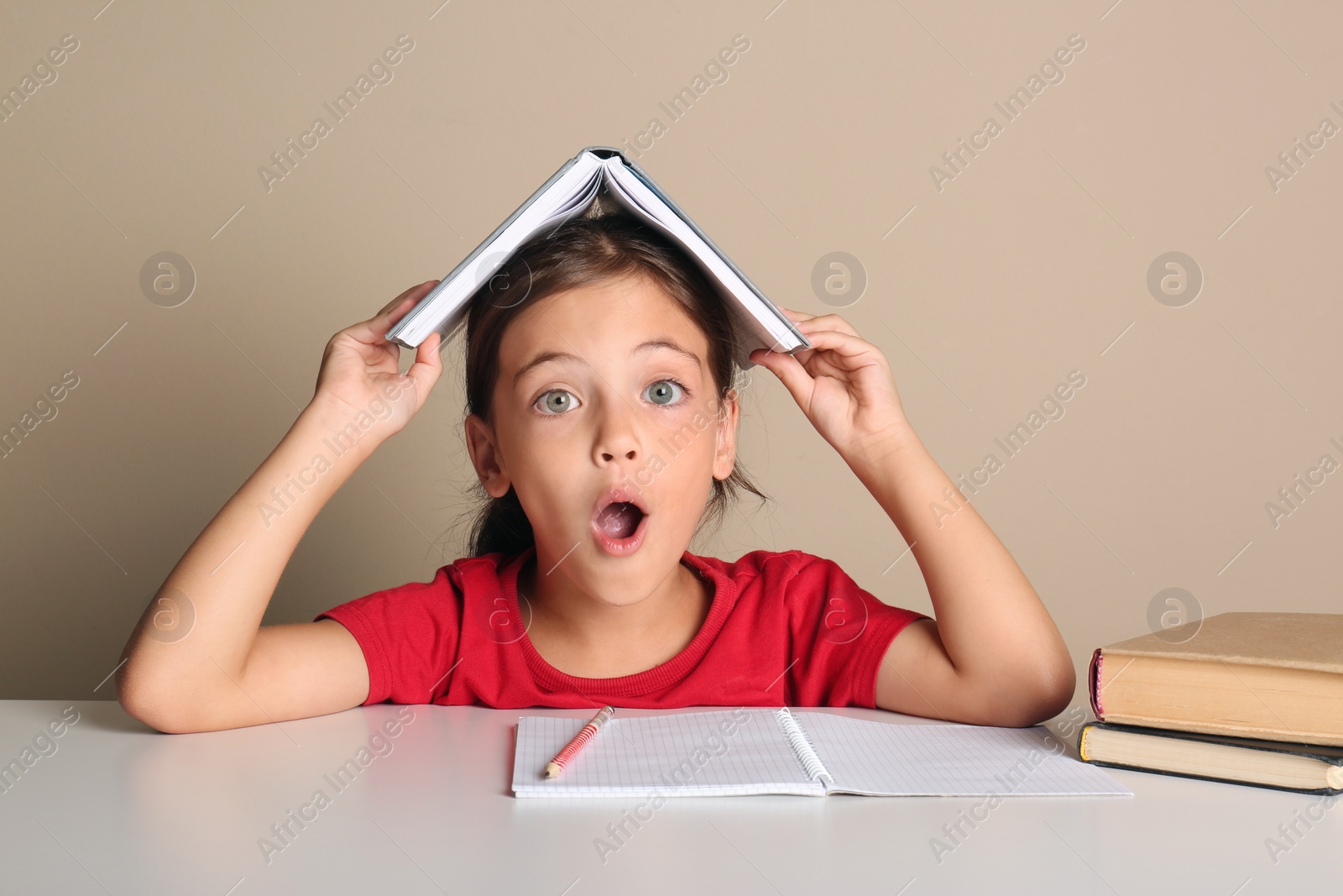 Photo of Emotional little girl with book on her head doing homework at table against beige background
