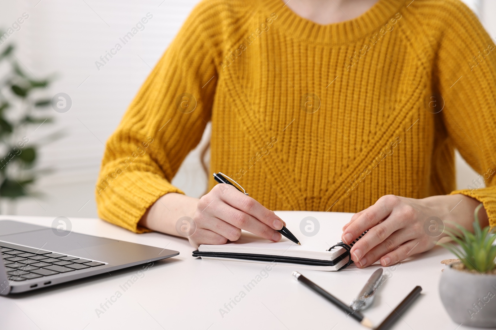 Photo of E-learning. Young woman taking notes during online lesson at white table indoors, closeup