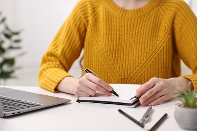 E-learning. Young woman taking notes during online lesson at white table indoors, closeup