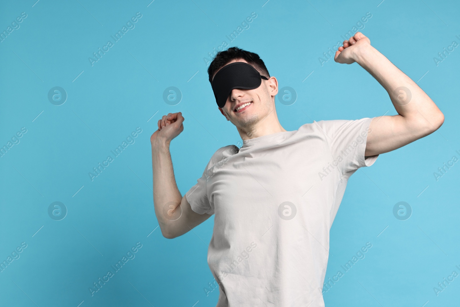 Photo of Happy man in pyjama and sleep mask stretching on light blue background, space for text