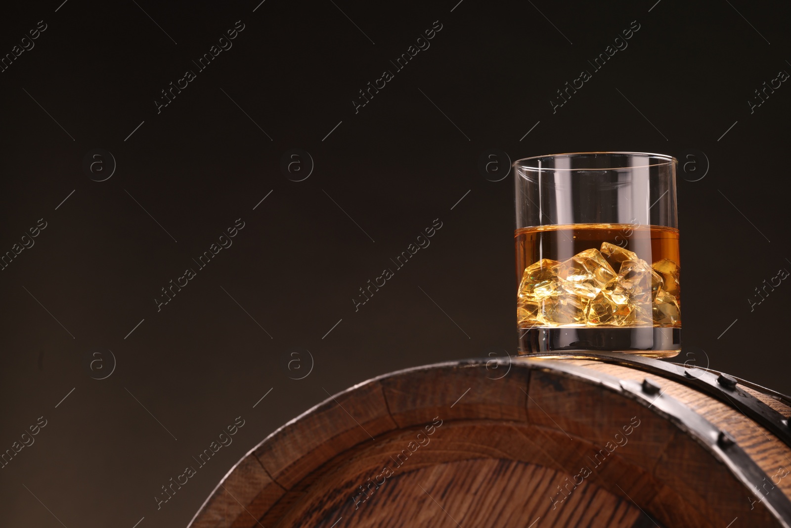 Photo of Whiskey with ice cubes in glass on wooden barrel against dark background, space for text