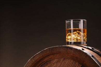 Whiskey with ice cubes in glass on wooden barrel against dark background, space for text