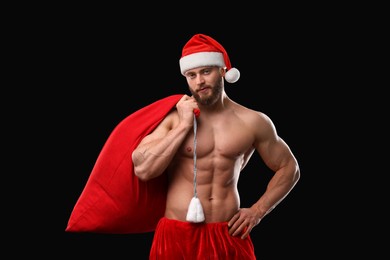 Muscular young man in Santa hat holding bag with presents on black background