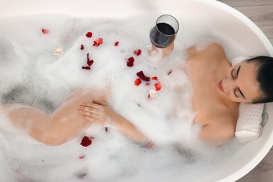 Photo of Woman with glass of wine taking bath in tub with foam and rose petals, top view