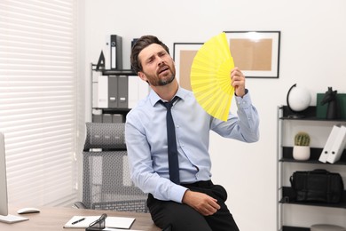 Bearded businessman waving yellow hand fan to cool himself in office