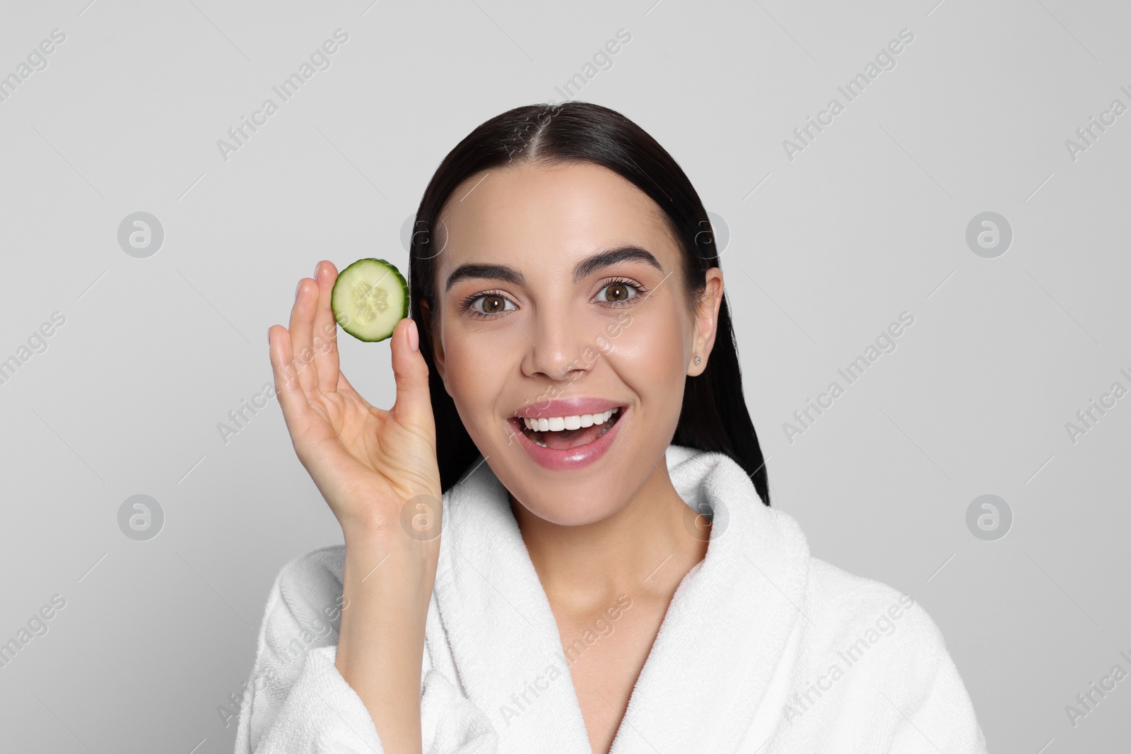 Photo of Woman in bathrobe holding piece of cucumber on light grey background. Spa treatment