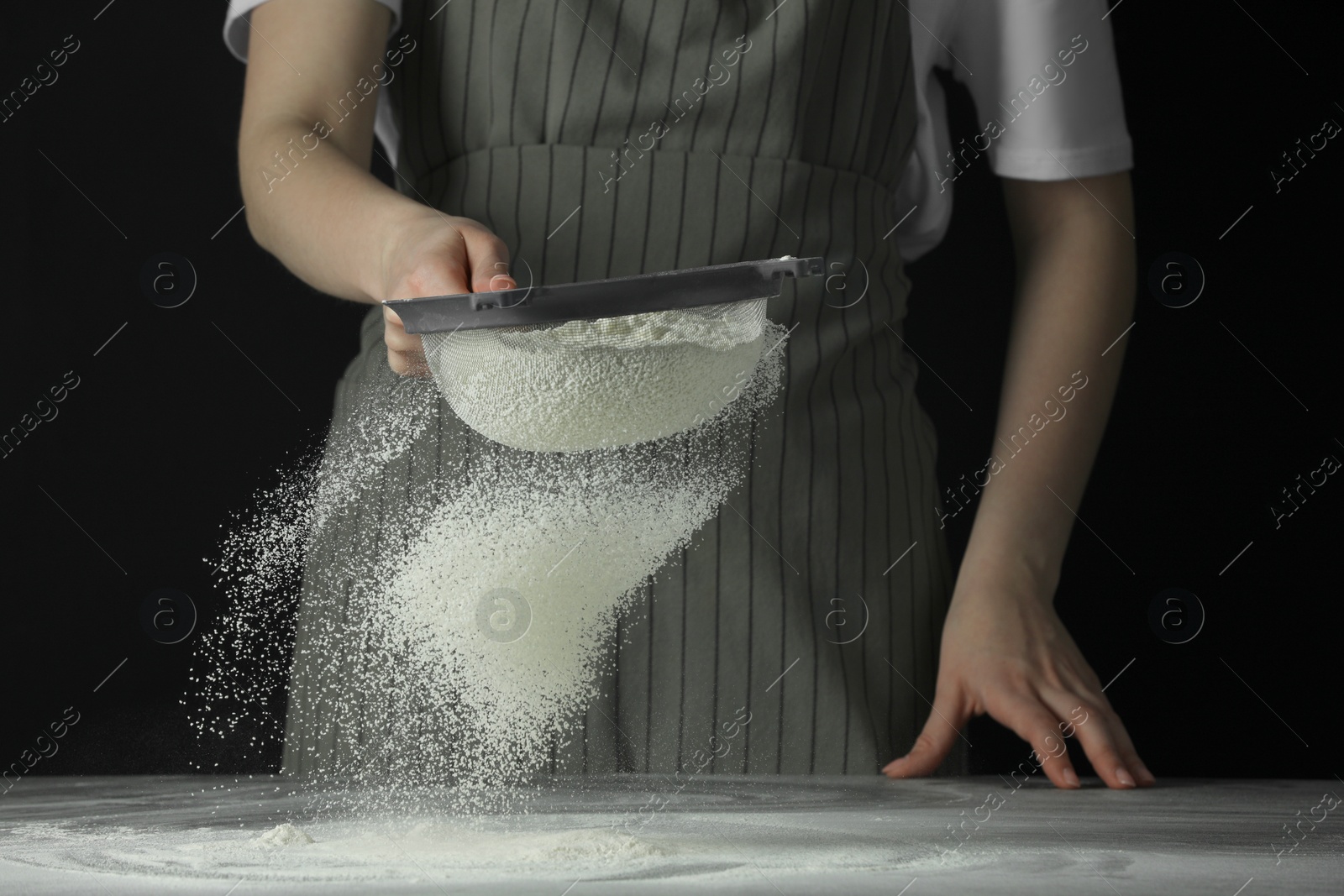 Photo of Woman sieving flour at table against black background, closeup