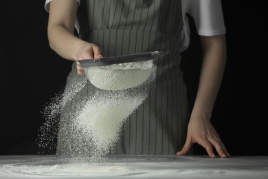 Woman sieving flour at table against black background, closeup