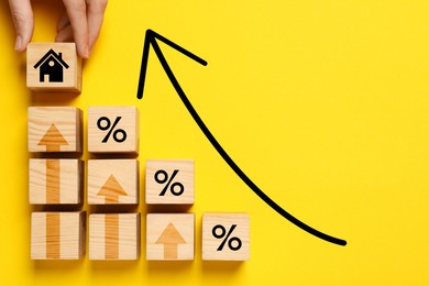 Image of Mortgage rate rising illustrated by upward arrows. Woman putting wooden cube with house icon near other ones on yellow background, top view