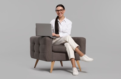 Happy woman with laptop sitting in armchair on light gray background