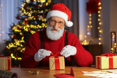 Photo of Santa Claus tying bow on gift box at his workplace in room decorated for Christmas