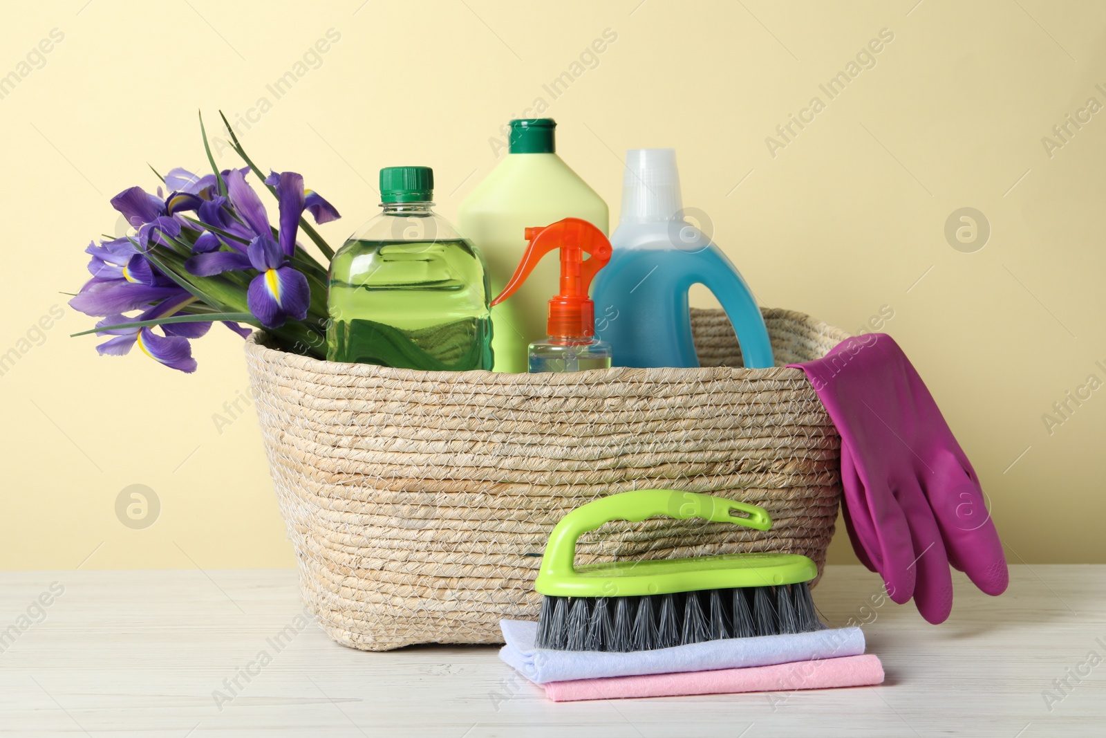 Photo of Spring cleaning. Wicker basket with detergents, flowers and gloves near tools on white wooden table
