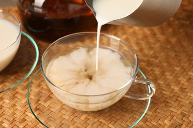 Photo of Pouring milk into glass cup at wicker mat, closeup