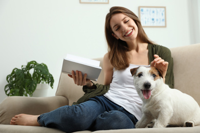 Photo of Young woman reading book and her cute Jack Russell Terrier on sofa at home. Lovely pet