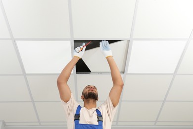 Electrician with screwdriver repairing ceiling light indoors, low angle view